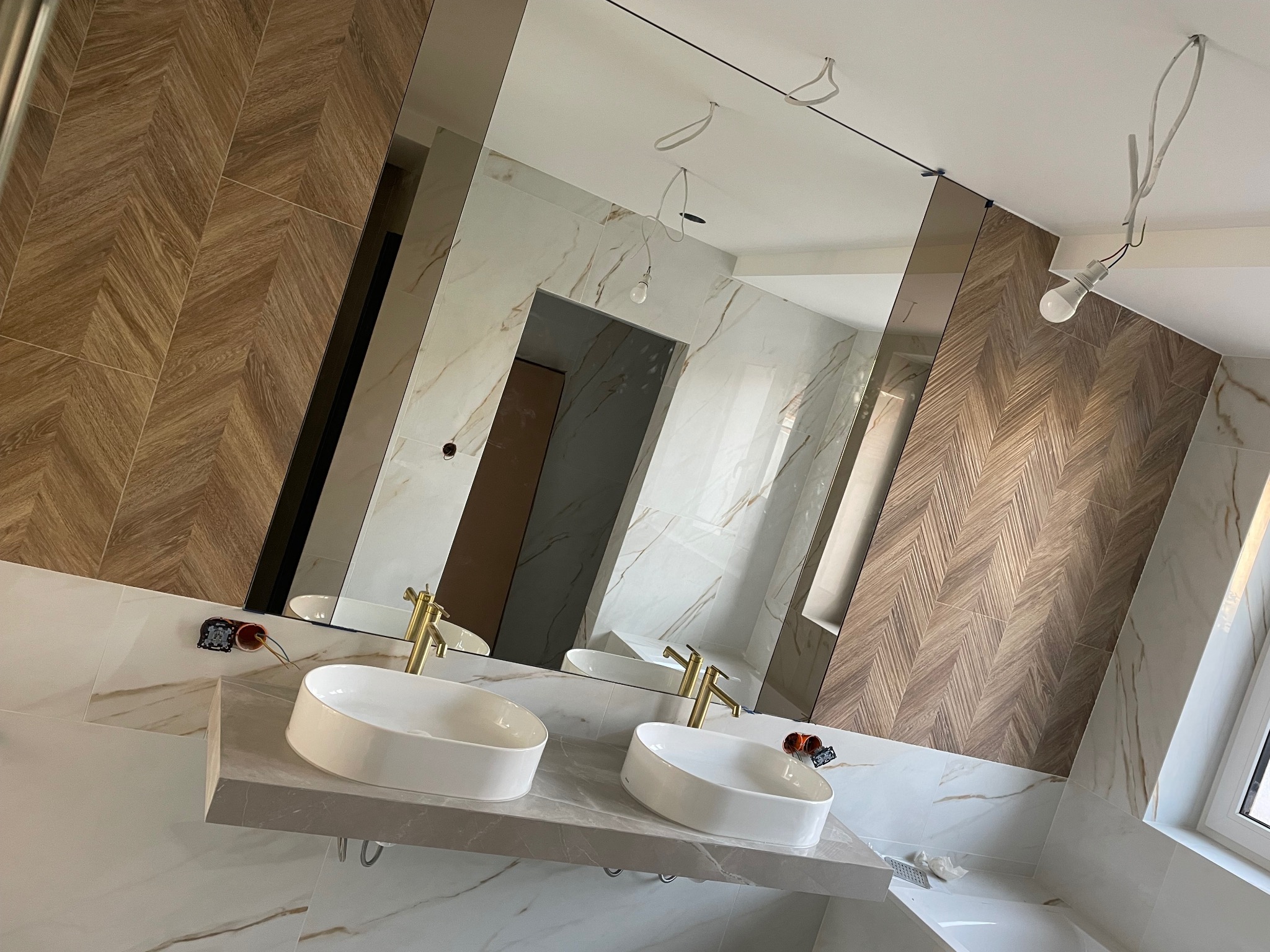 Wykończona łazienka z dwoma umywalkami nablatowymi, złotą armaturą i lustrem na ścianie. Na ścianach płytki imitujące drewno ułożone w jodełkę i marmur z żyłami.