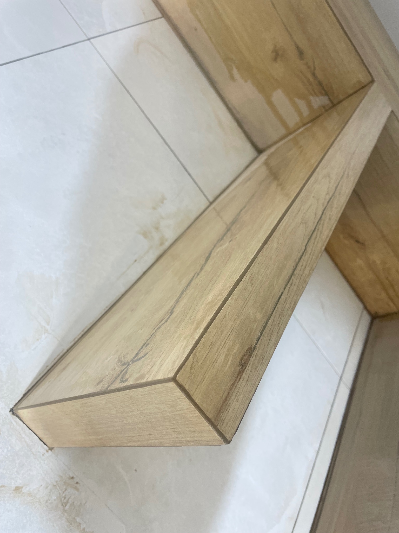 Wykończona półka z płytek imitujących drewno, wkomponowana w białe, błyszczące płytki ścienne. Detal wykończenia wnętrza z bliska, z widocznymi fugami.