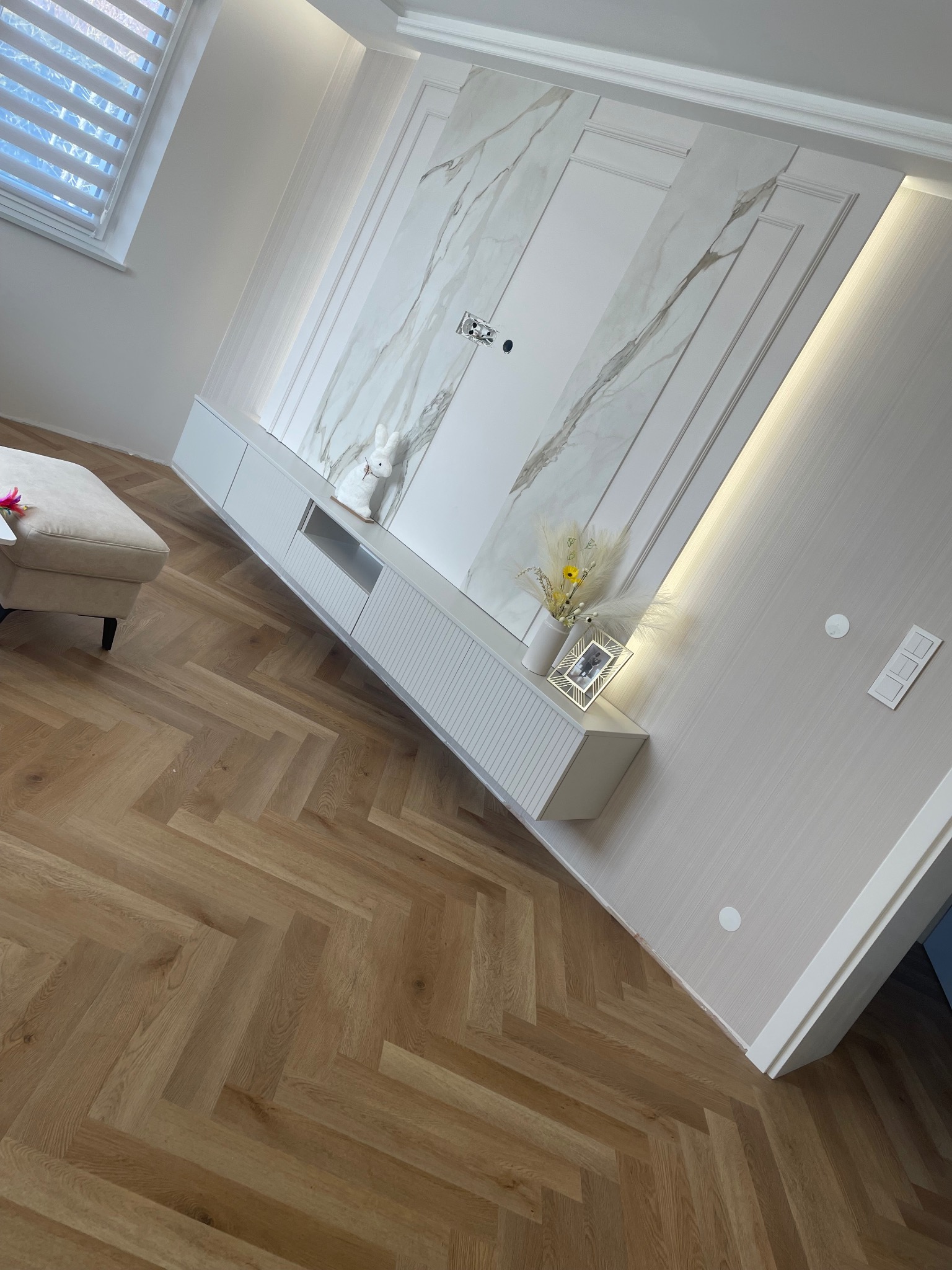 Eleganckie wykończenie wnętrza: parkiet w jodełkę, ściana z imitacją marmuru, biała zabudowa meblowa z dekoracjami i subtelnym oświetleniem.