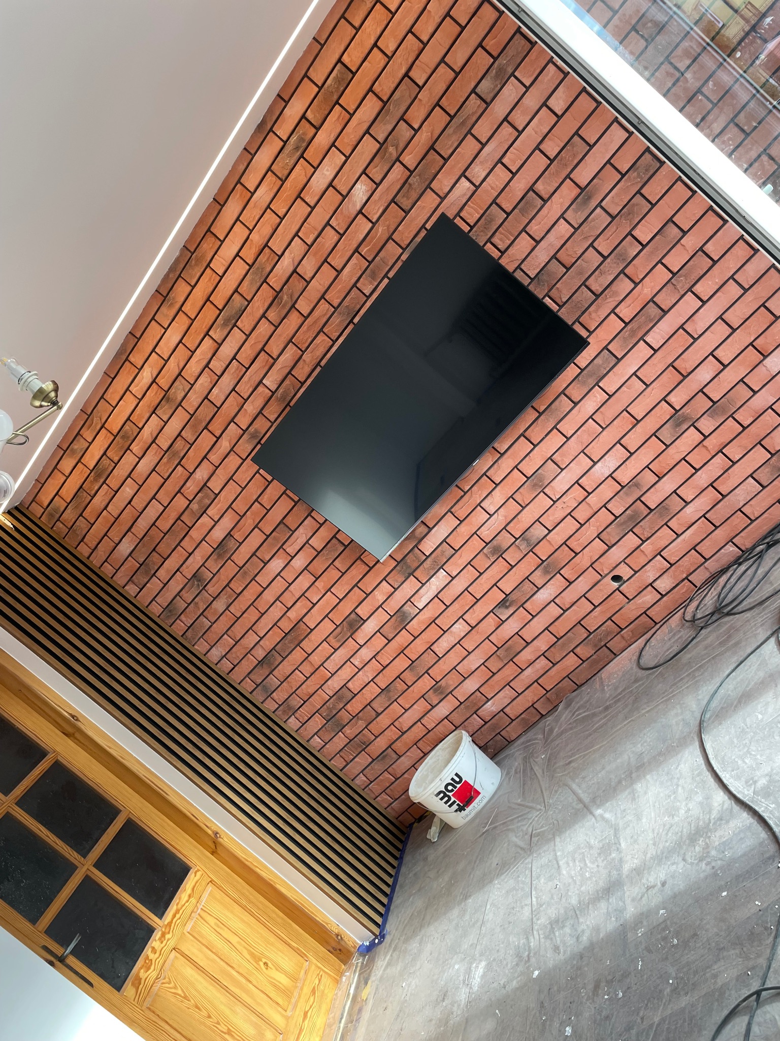 Ściana z cegły klinkierowej z wnęką na TV i panelami dekoracyjnymi w Turobinie. Widoczny fragment drzwi i wiadro z farbą. Nietypowy kąt ujęcia.