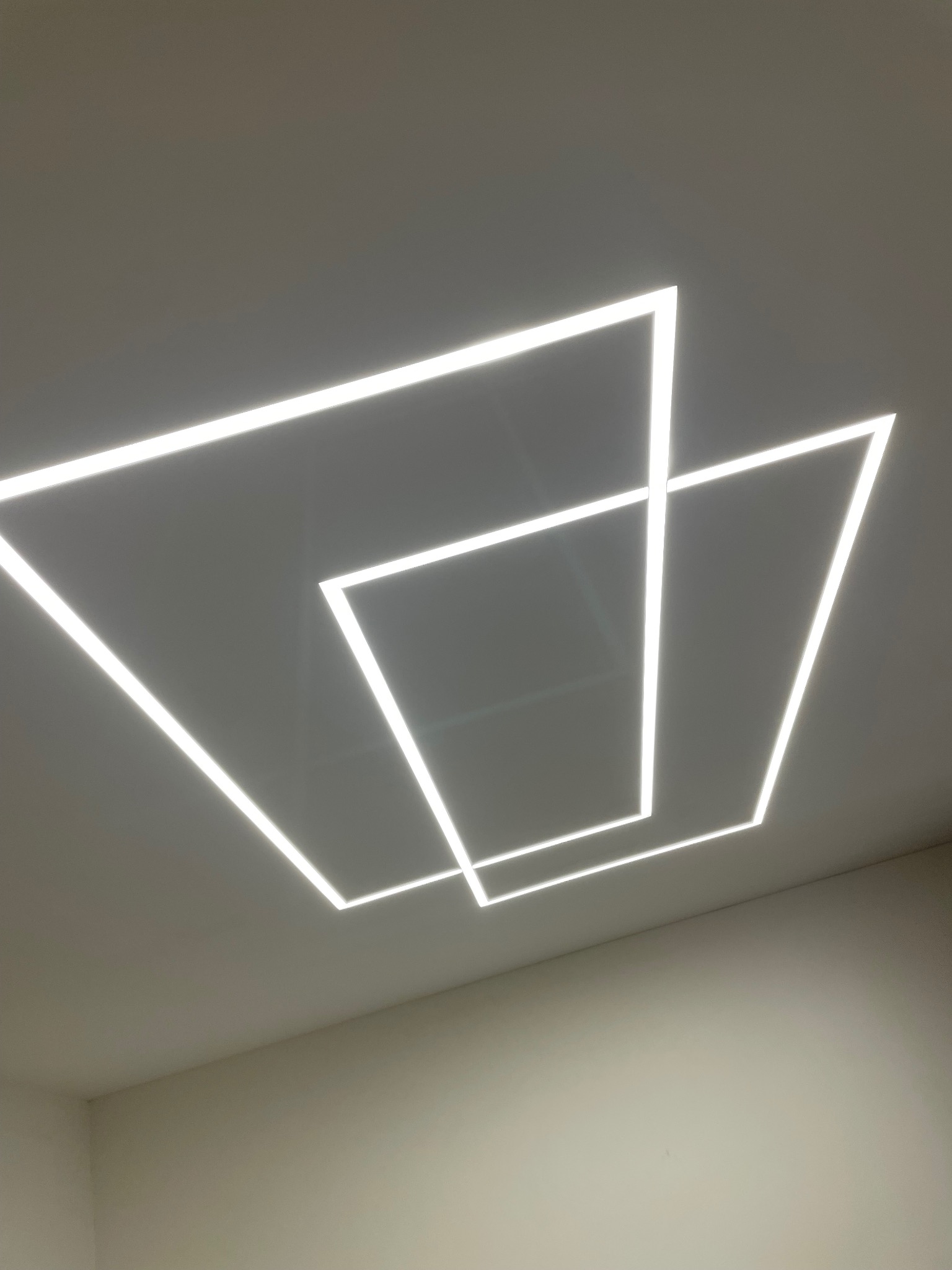 Nowoczesne oświetlenie sufitowe LED w kształcie nakładających się kwadratów, emitujące jasne, białe światło w minimalistycznym wnętrzu.