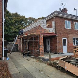 Jacek Kasprzycki - Rozbudowa domu z cegły: murarz na rusztowaniu, wznoszenie ścian z bloczków. Widoczne materiały budowlane i elementy konstrukcyjne.