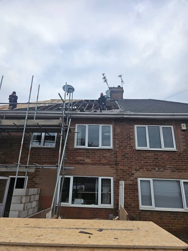 Dwóch robotników na dachu szeregowca podczas remontu. Widoczne rusztowania, częściowo zdjęte pokrycie dachowe i antena satelitarna. Przebudowa dachu w trakcie.