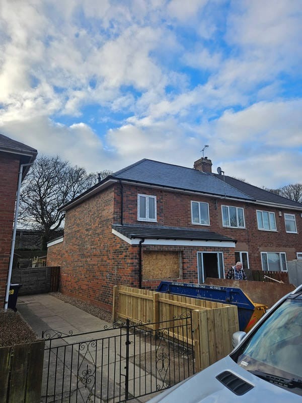 Dwupiętrowy dom z cegły po remoncie dachu, z nową elewacją i ogrodzeniem. Kontener na gruz przed budynkiem. Widok z zewnątrz, pochmurne niebo.