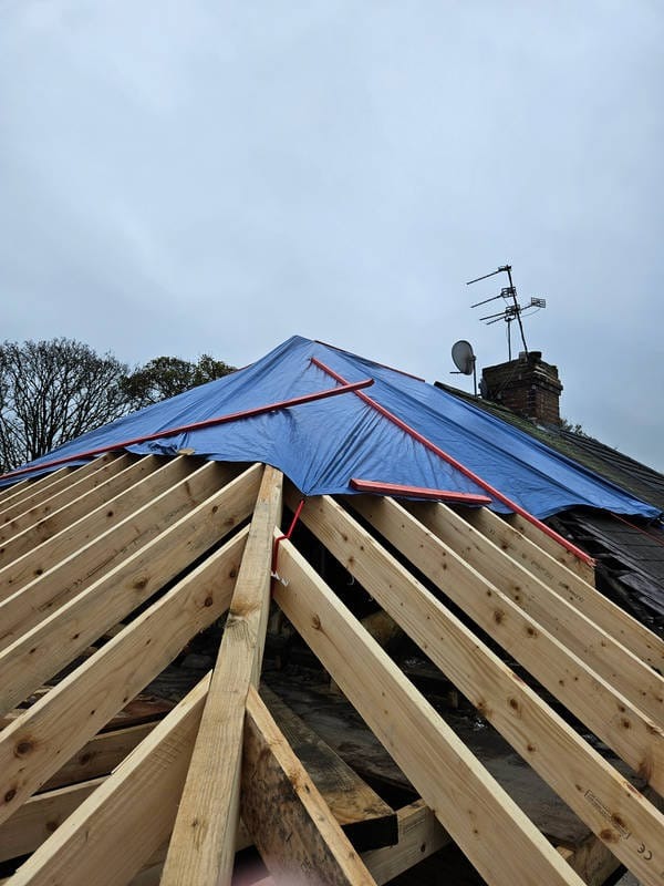 Konstrukcja dachu z drewnianych belek, zabezpieczona niebieską plandeką z czerwonymi elementami mocującymi, na tle pochmurnego nieba i komina z anteną.