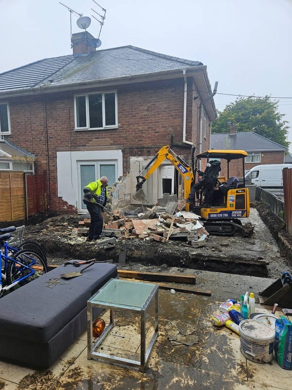 Demolka ściany domu z cegły przy użyciu koparki, widoczny pracownik w odblaskowej kamizelce. Na pierwszym planie meble i narzędzia.