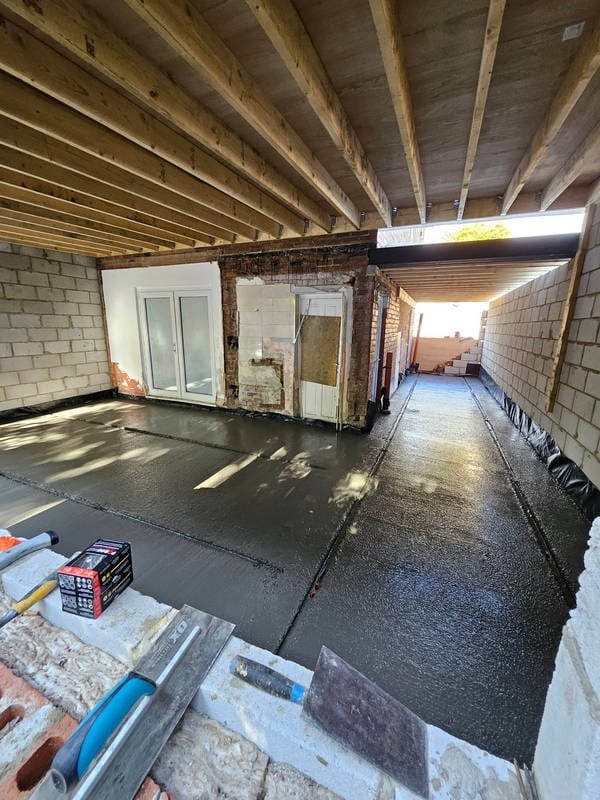 Wylana, mokra posadzka w surowym wnętrzu z odsłoniętymi cegłami i drewnianą konstrukcją stropu. Widoczne narzędzia murarskie i poziomica. Etap prac wykończeniowych.