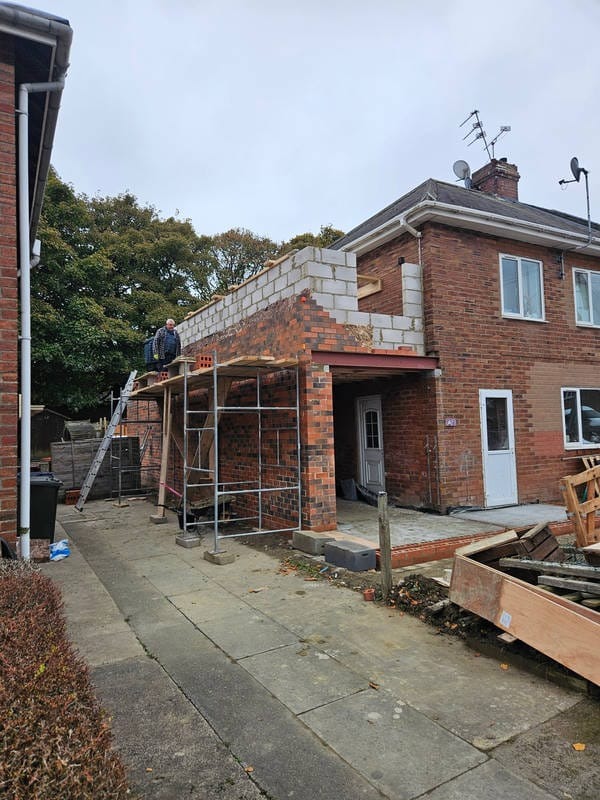 Rozbudowa domu z cegły: murarz na rusztowaniu, wznoszenie ścian z bloczków. Widoczne materiały budowlane i elementy konstrukcyjne.