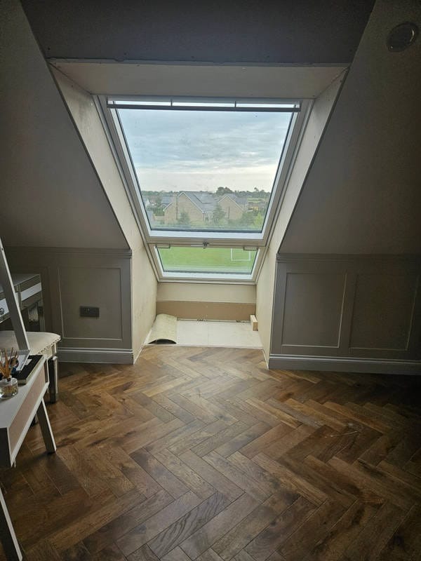 Podłoga z parkietu ułożonego w jodełkę w pokoju na poddaszu z oknem dachowym. Widoczne fragmenty ścian z dekoracyjnymi panelami i przygotowane podłoże pod oknem.