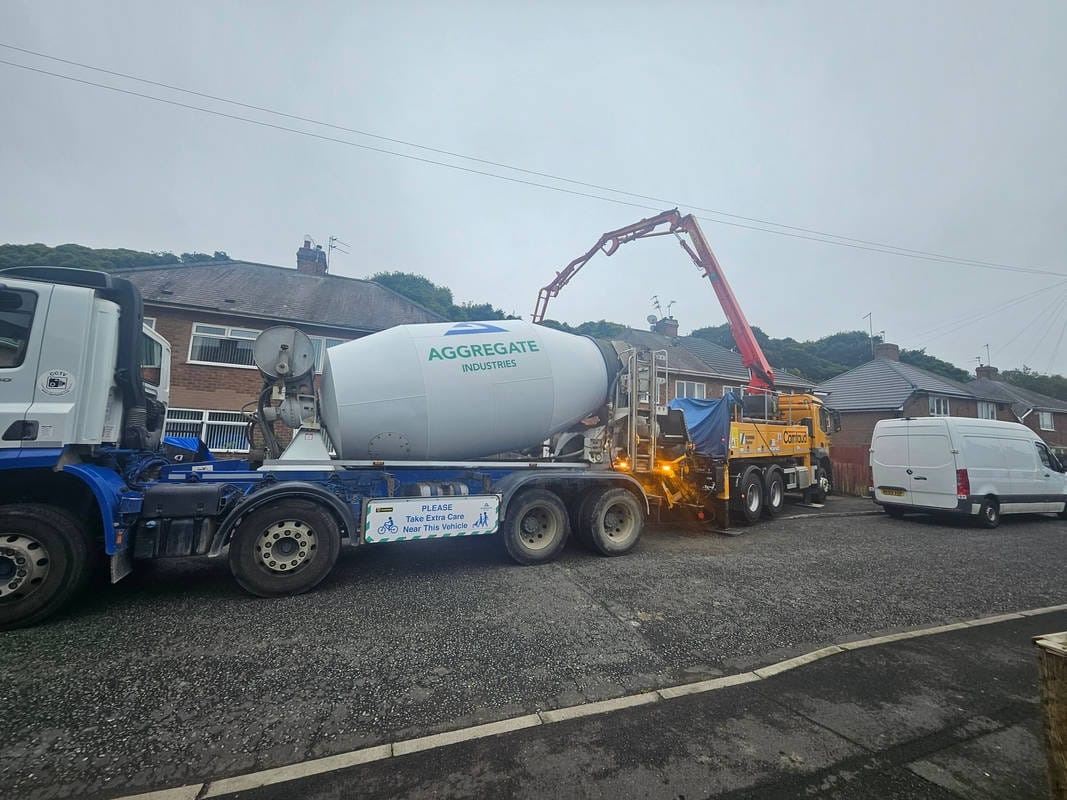 Ciężarówka z betonomieszarką Aggregate Industries z wysięgnikiem pompy do betonu na tle domów jednorodzinnych w pochmurny dzień. Tabliczka ostrzegawcza na pojeździe.