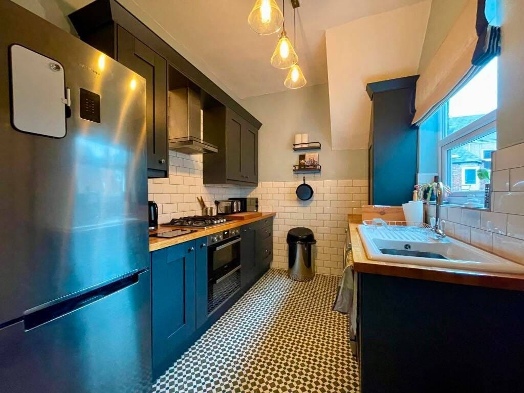 Niebieska kuchnia z białymi płytkami i wzorzystą podłogą. Stalowa lodówka Samsung, drewniany blat i wiszące lampy dodają elegancji. Widok na okno z roletą.