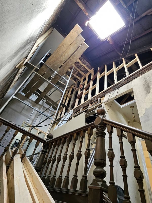 Wnętrze domu w trakcie remontu: drewniane schody z balustradą, rusztowania, odsłonięta konstrukcja dachu z oknem dachowym, widoczny pracownik na rusztowaniu.