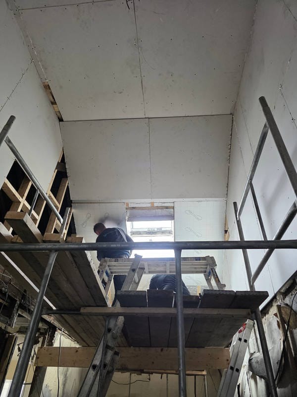 Montaż płyt gipsowo-kartonowych na suficie poddasza, widok z rusztowania, okno dachowe w tle, surowe wykończenie ścian.