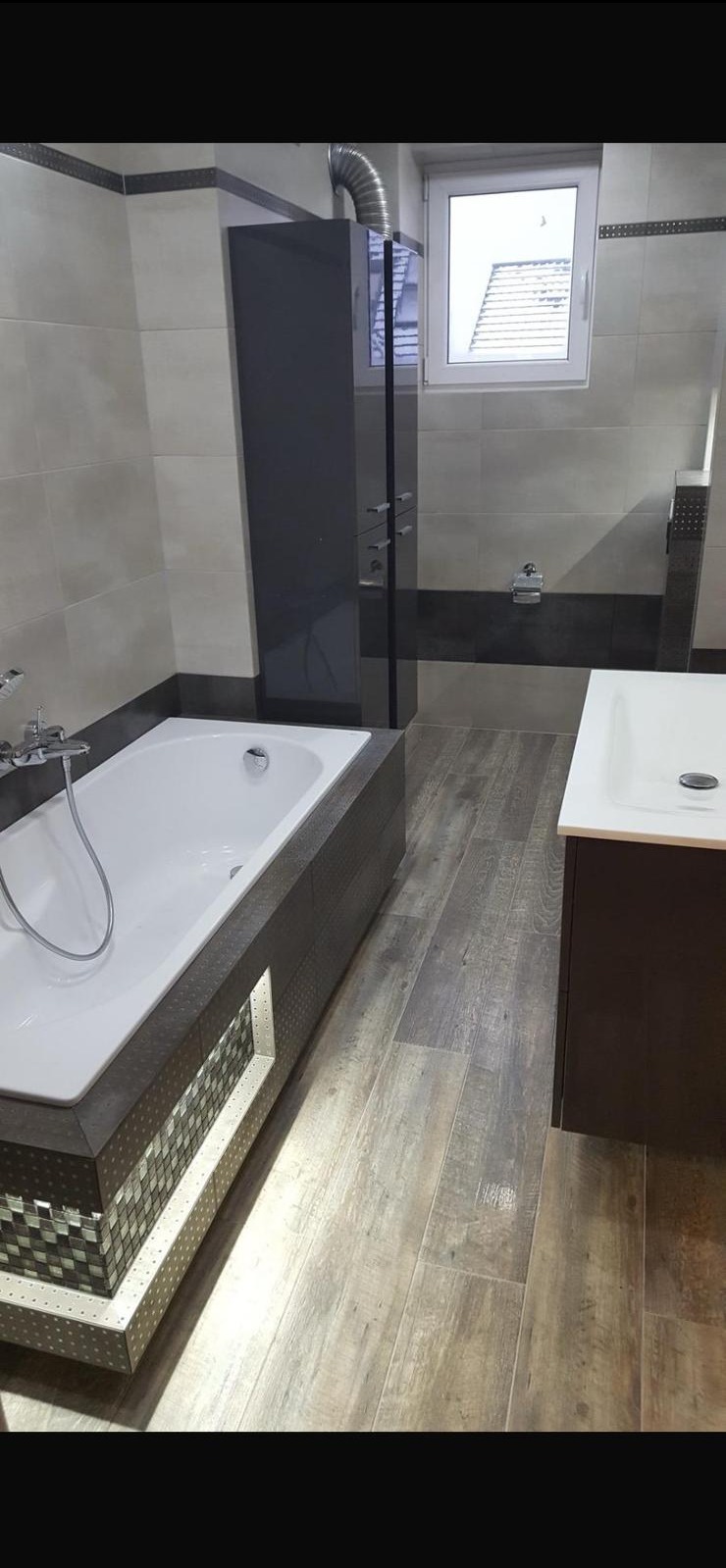 Nowoczesna łazienka z wanną obudowaną mozaiką i podświetleniem LED, drewnopodobne płytki na podłodze, szara szafka z umywalką i kabina prysznicowa.