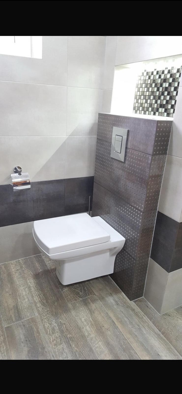 Nowoczesna toaleta podwieszana w wyremontowanej łazience w Hessisch Lichtenau. Szare płytki z perforacją, drewnopodobna podłoga, chromowany uchwyt na papier.