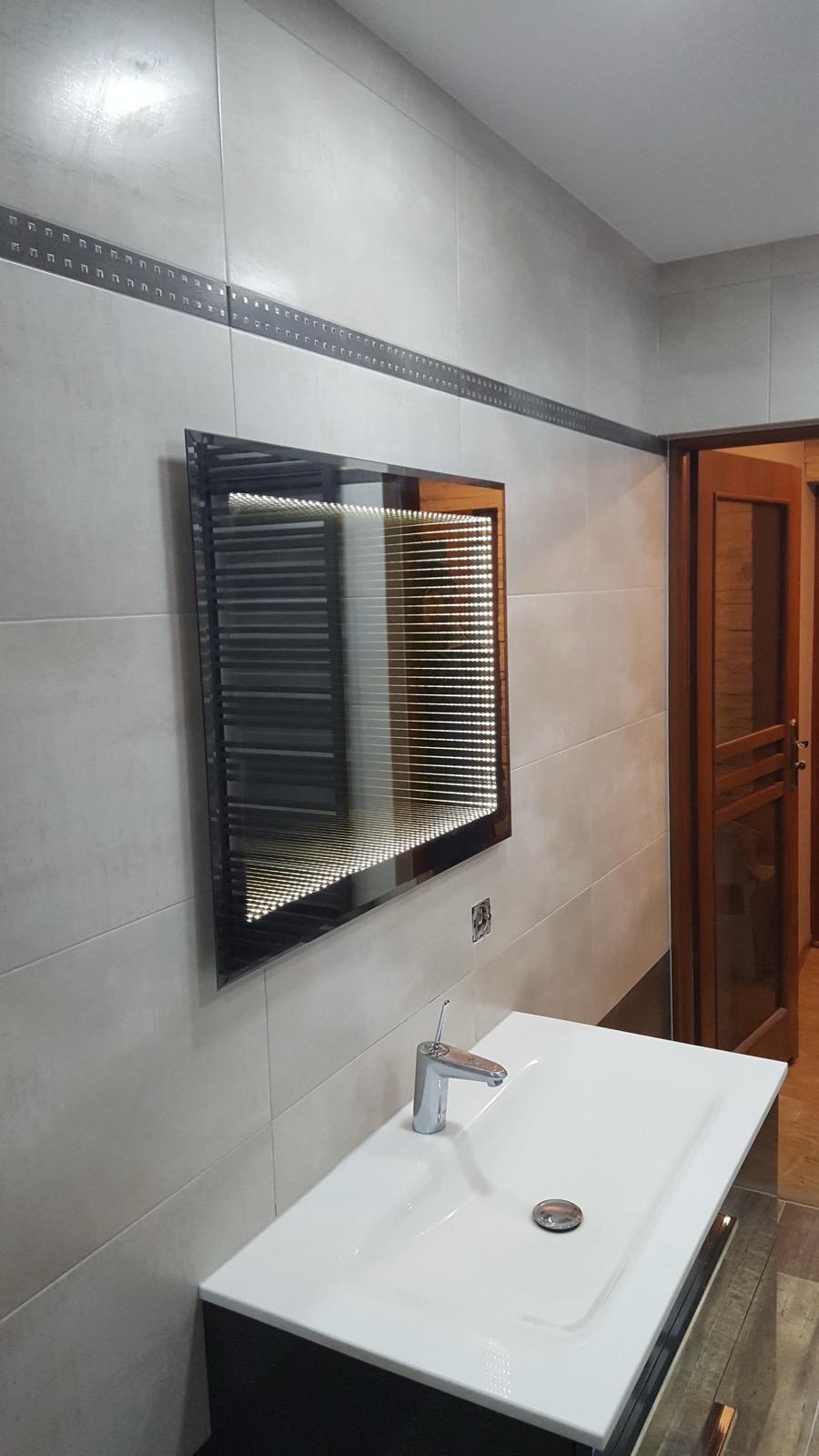 Wykończona łazienka z umywalką i lustrem nieskończoności. Płytki w odcieniach beżu z dekoracyjnym paskiem. Widoczne drewniane drzwi i podłoga.