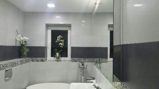 Nowoczesna łazienka w bieli i szarości z akcentem mozaiki, kwiatami w oknie i dużym lustrem. Minimalistyczny design i eleganckie wykończenie.