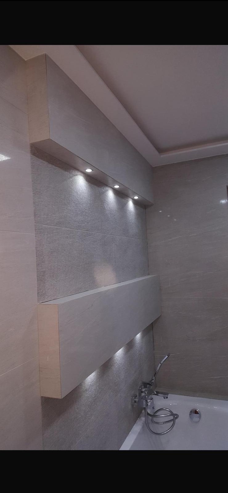 Wykończona łazienka z szarymi płytkami, podświetlanymi półkami i wanną. Minimalistyczny design z subtelnym oświetleniem punktowym.