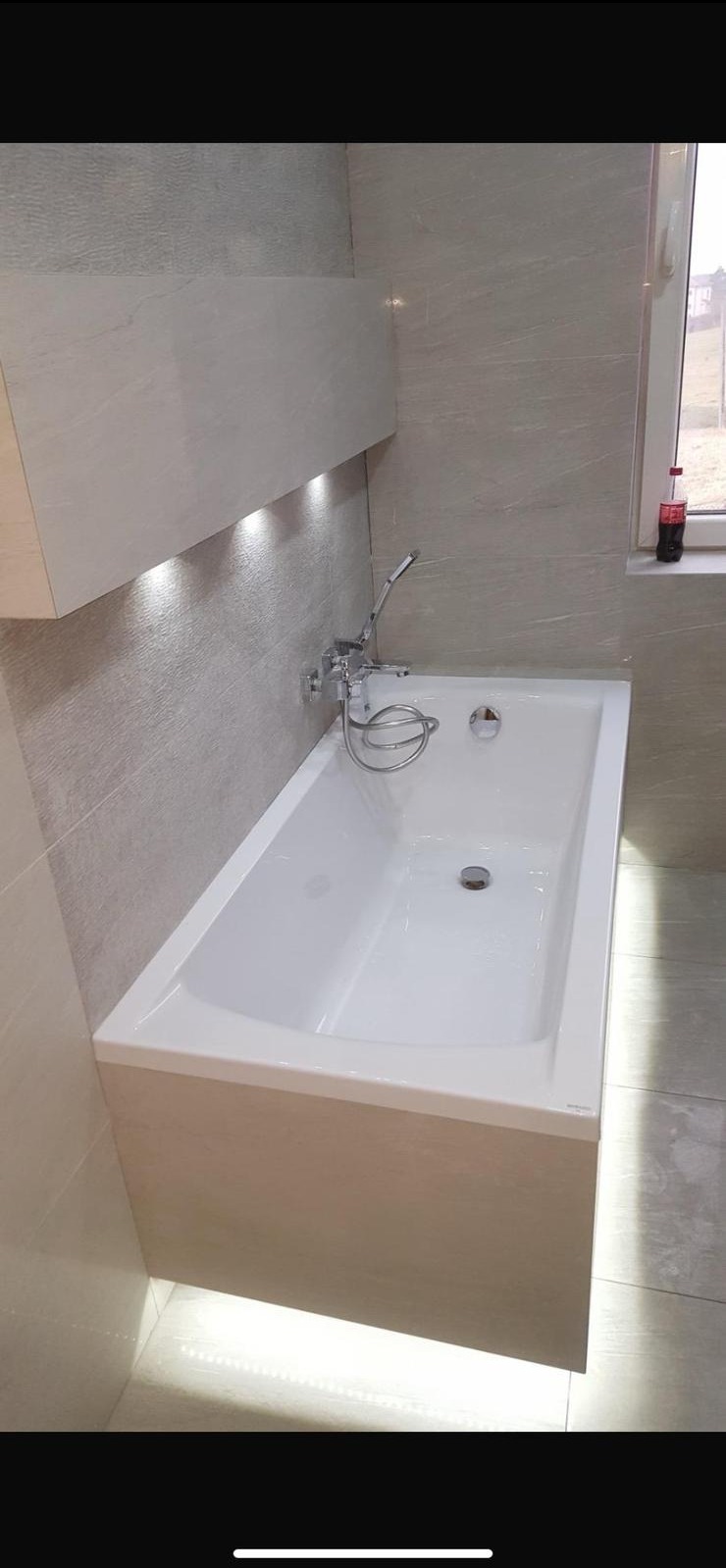 Biała wanna z chromowaną armaturą w wykończonej łazience z beżowymi płytkami i podświetleniem LED. Na parapecie stoi butelka Coca-Coli.