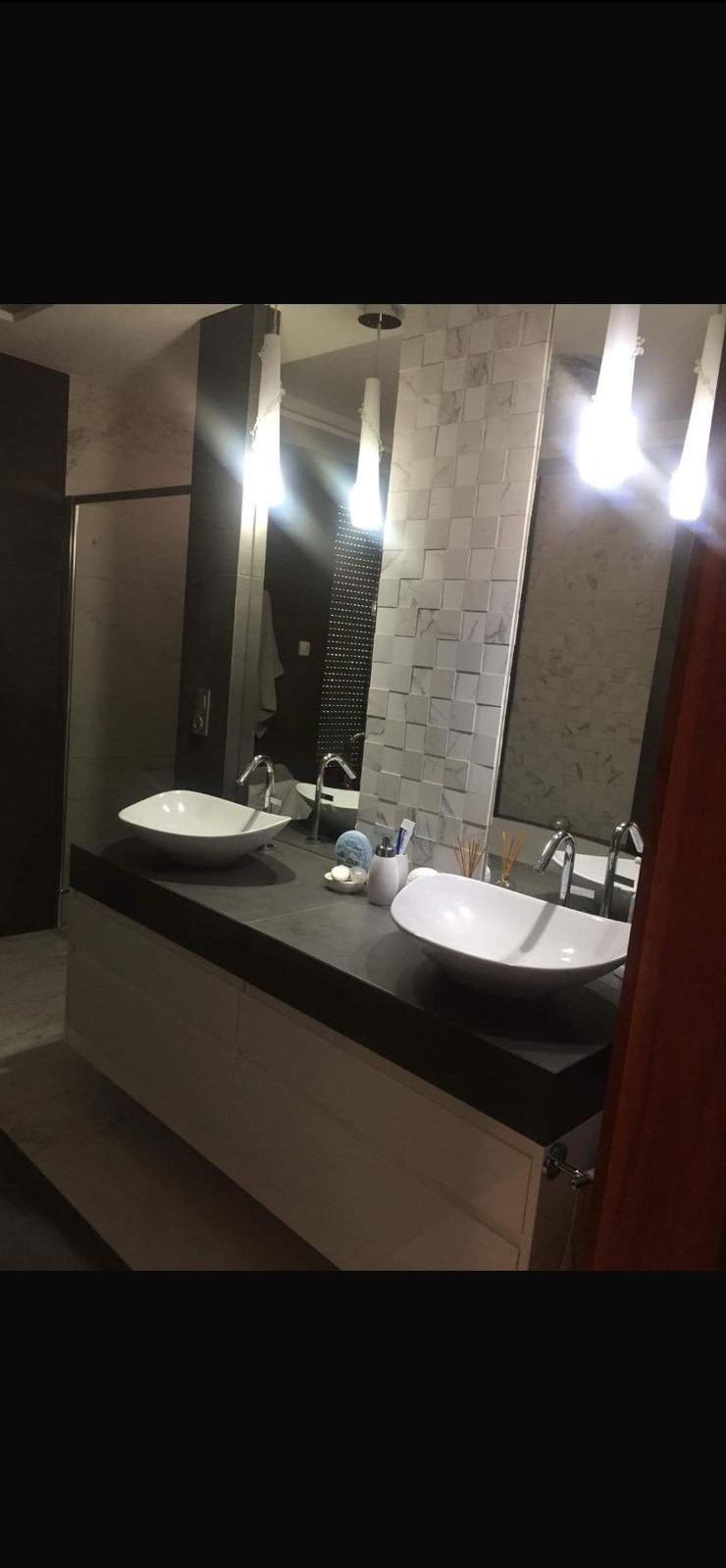 Nowoczesna łazienka z dwoma umywalkami w kształcie łezki, ciemnym blatem i białymi szafkami. Na ścianie dekoracyjne płytki 3D i podłużne lampy nad lustrami.