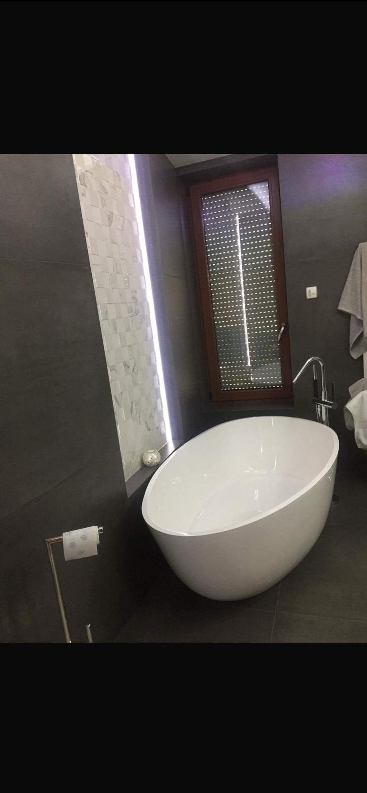 Nowoczesna łazienka z wolnostojącą wanną o owalnym kształcie, szarą ścianą i oknem z roletami. Akcent stanowi podświetlana ściana z mozaiką.