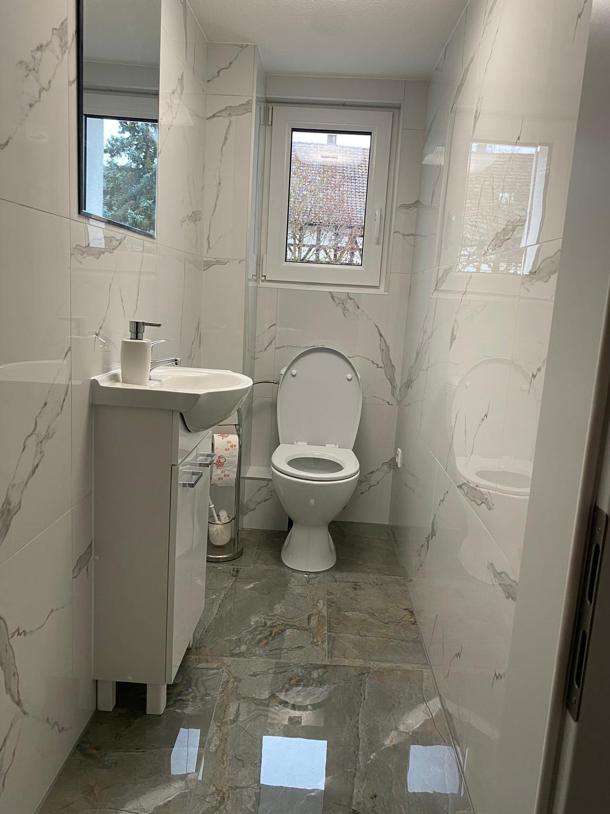 Biała łazienka z marmurowymi płytkami na ścianach i szarymi płytkami na podłodze. Umywalka z szafką, toaleta i okno. Widoczne odbicie w płytkach podłogowych.