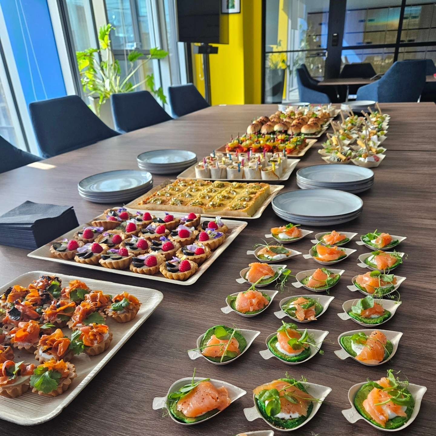 Stół konferencyjny z różnorodnymi przekąskami: tartaletki z owocami, mini-burgery, roladki i kanapeczki z łososiem. Idealne na firmowe spotkanie lub event.