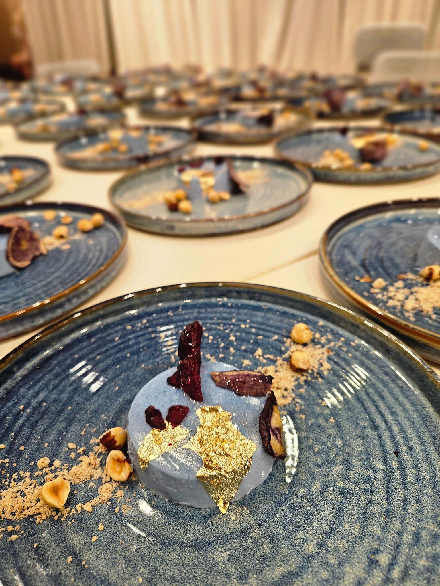 Elegancko podane desery na niebieskich talerzach z orzechami i złotymi płatkami, ustawione rzędami na białym stole. Dekoracyjne i wyrafinowane słodkości.