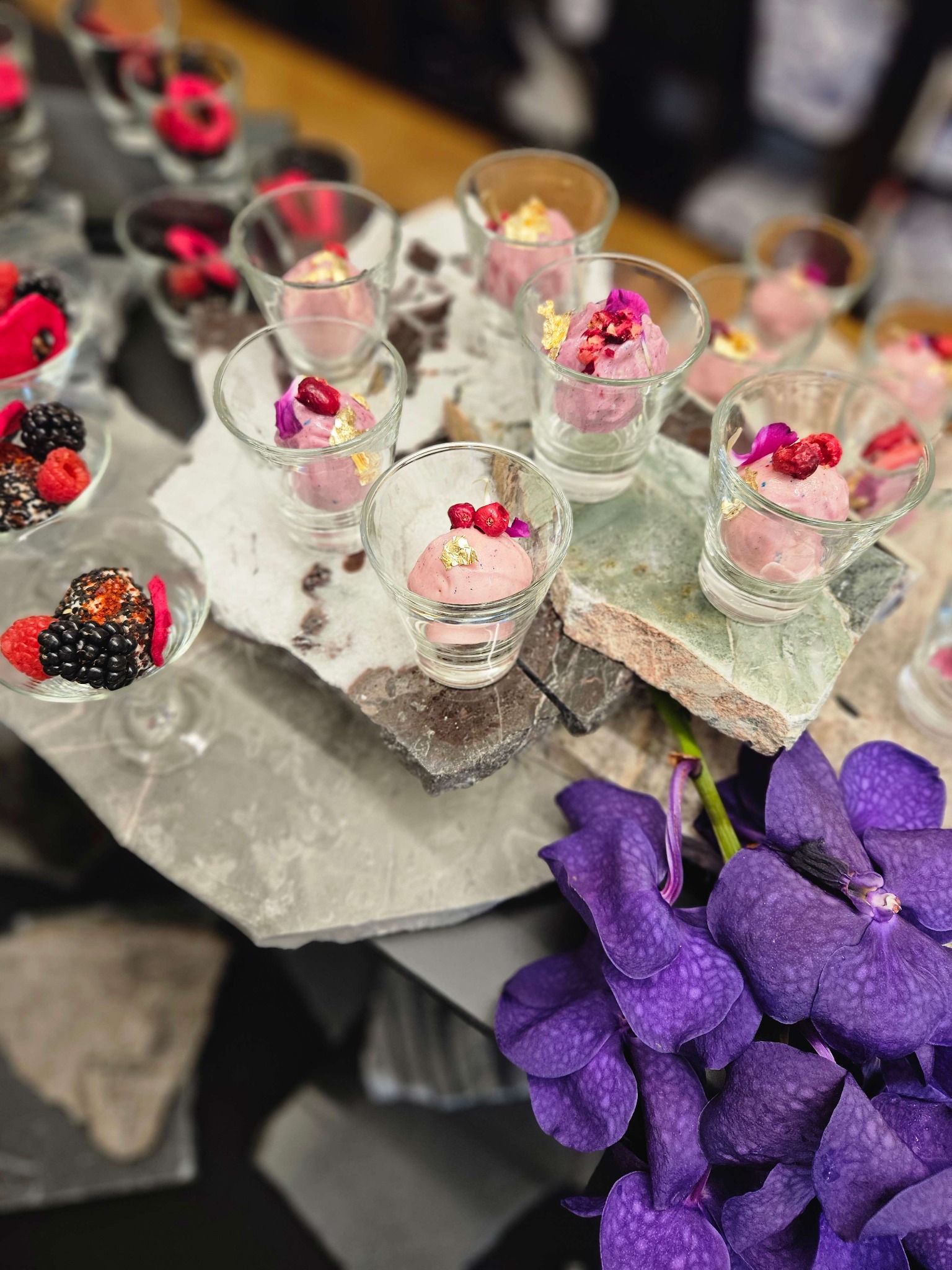 Deser lodowy w szklankach, udekorowany owocami i płatkami złota, na kamiennej płycie z fioletowymi kwiatami. Elegancki catering.