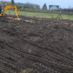 TerraTech - Żółta koparka na polu z przygotowaną ziemią, widoczne ślady opon. W tle ogrodzenie z drewnianych słupków i budynek mieszkalny. Pochmurne niebo.