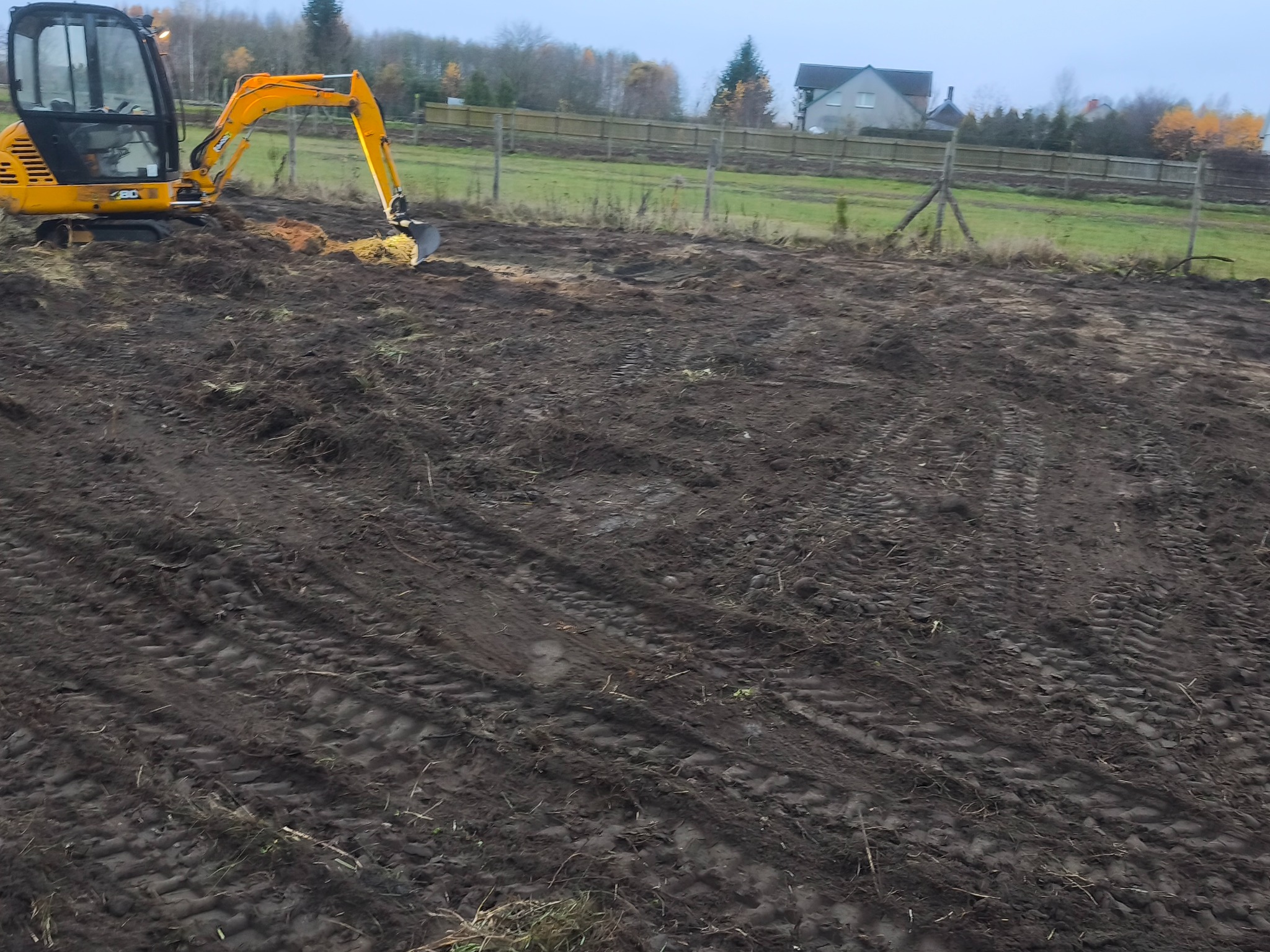 Żółta koparka na polu z przygotowaną ziemią, widoczne ślady opon. W tle ogrodzenie z drewnianych słupków i budynek mieszkalny. Pochmurne niebo.