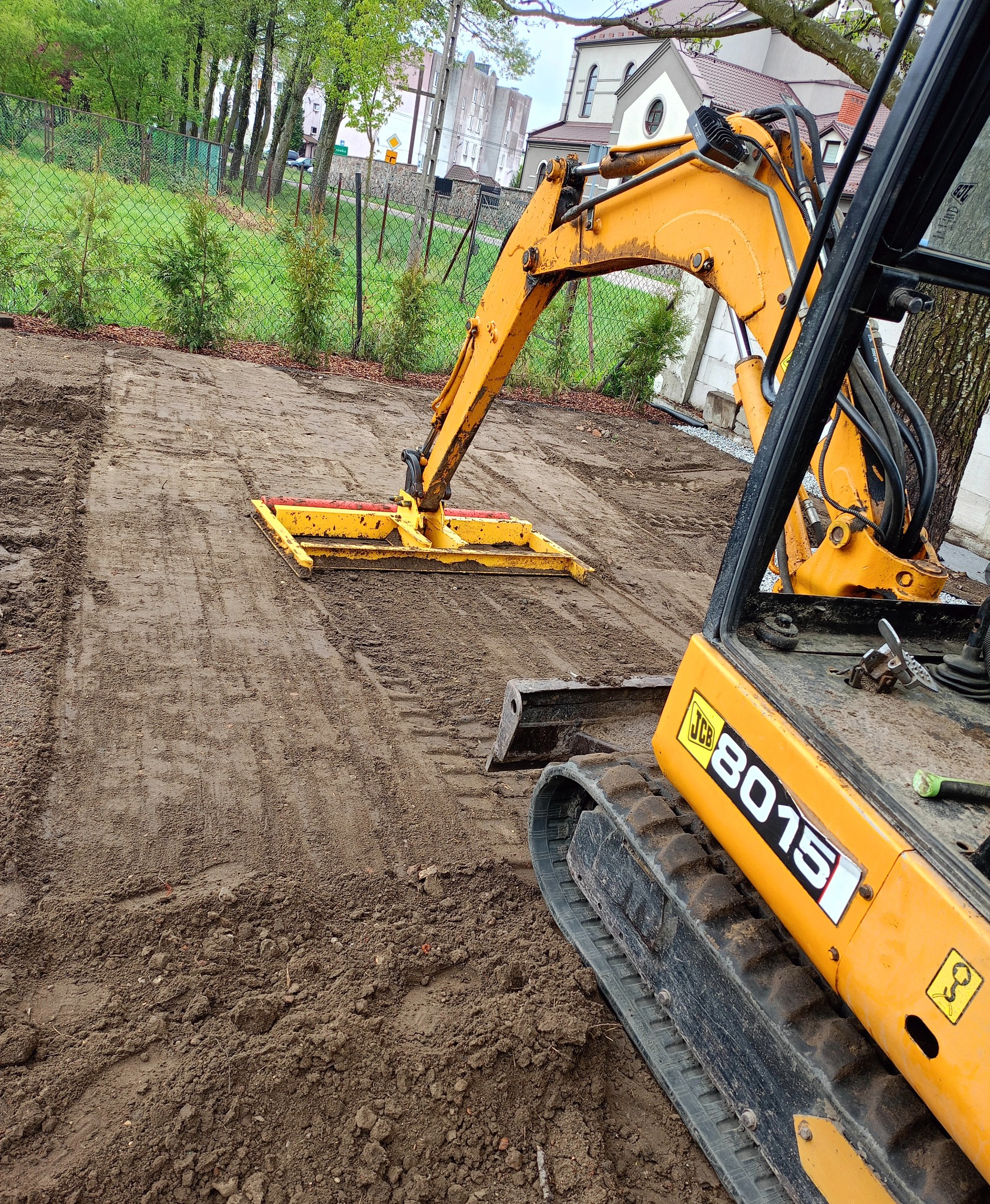Żółta minikoparka JCB 8015 na placu budowy, przygotowuje teren pod inwestycję. Widoczne ślady opon na ziemi i fragment budynku w tle.