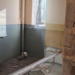 MARCIN MICAŁ - Wnętrze pomieszczenia w trakcie rozbiórki: odsłonięte rury, fragmenty gruzu na podłodze, ślady po usuniętych płytkach. Widoczne elementy instalacji i fragment okna z firanką.