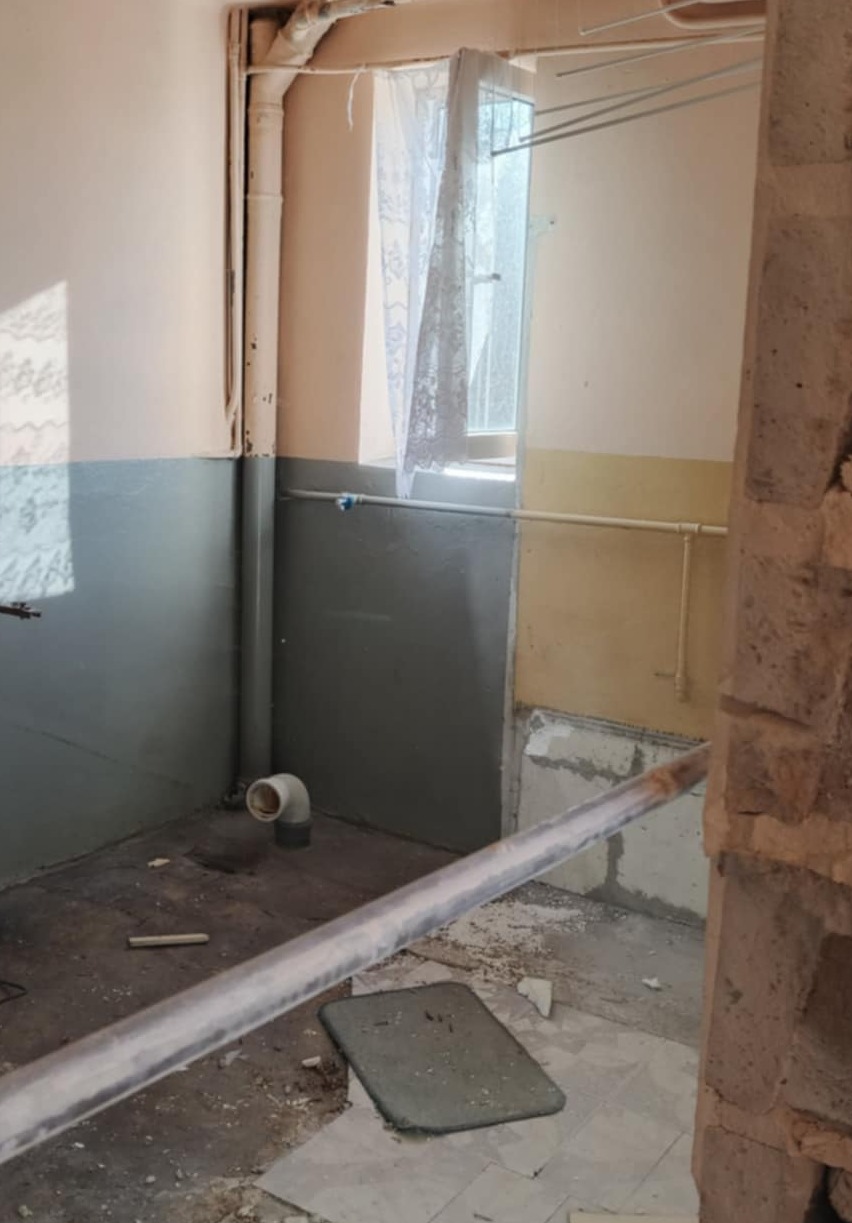 Wnętrze pomieszczenia w trakcie rozbiórki: odsłonięte rury, fragmenty gruzu na podłodze, ślady po usuniętych płytkach. Widoczne elementy instalacji i fragment okna z firanką.