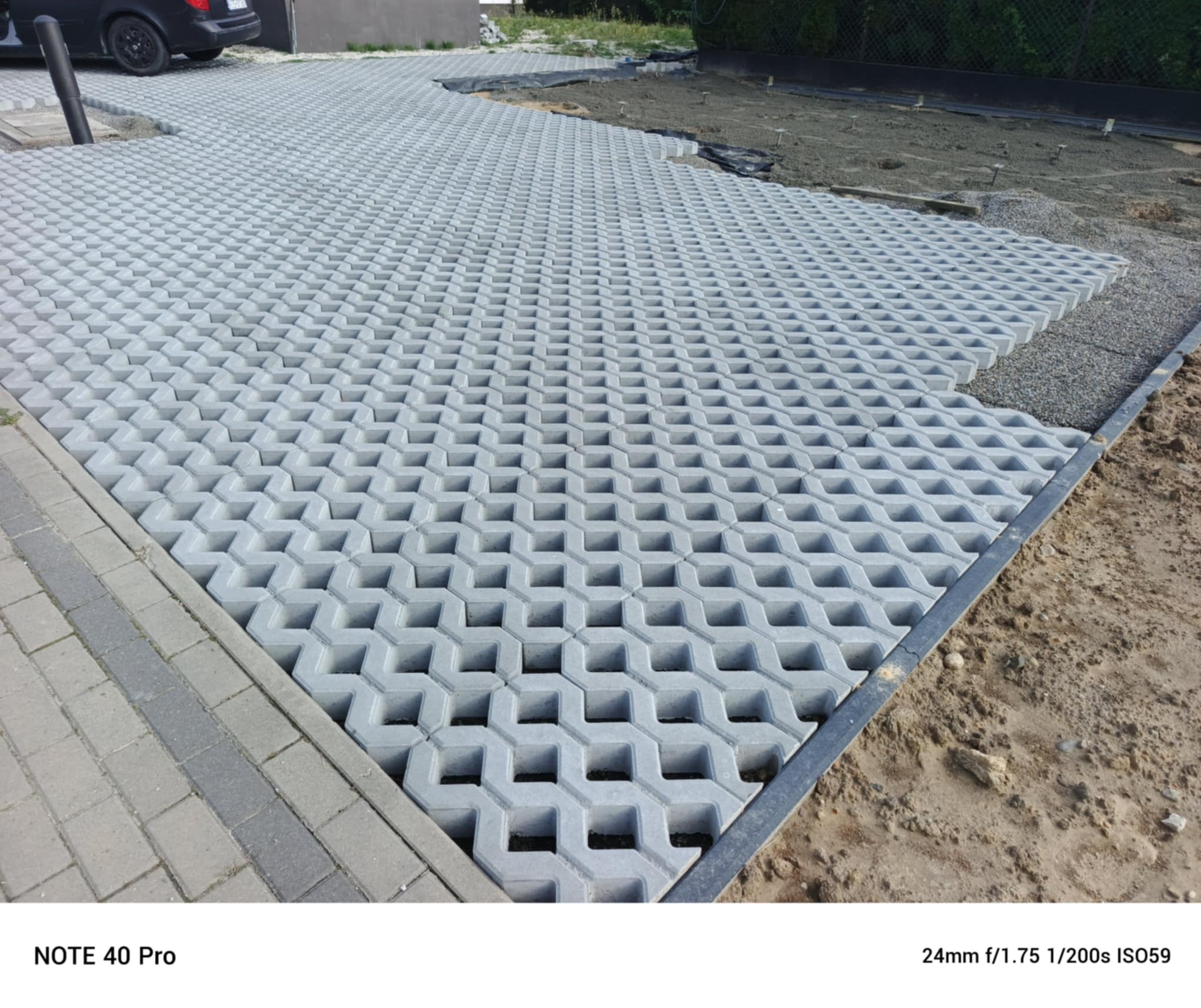 Szara, ażurowa kostka brukowa Eko ułożona na podjeździe, widok z góry. Częściowo zasypana żwirem. W tle zaparkowany samochód i teren przygotowany pod dalsze prace.