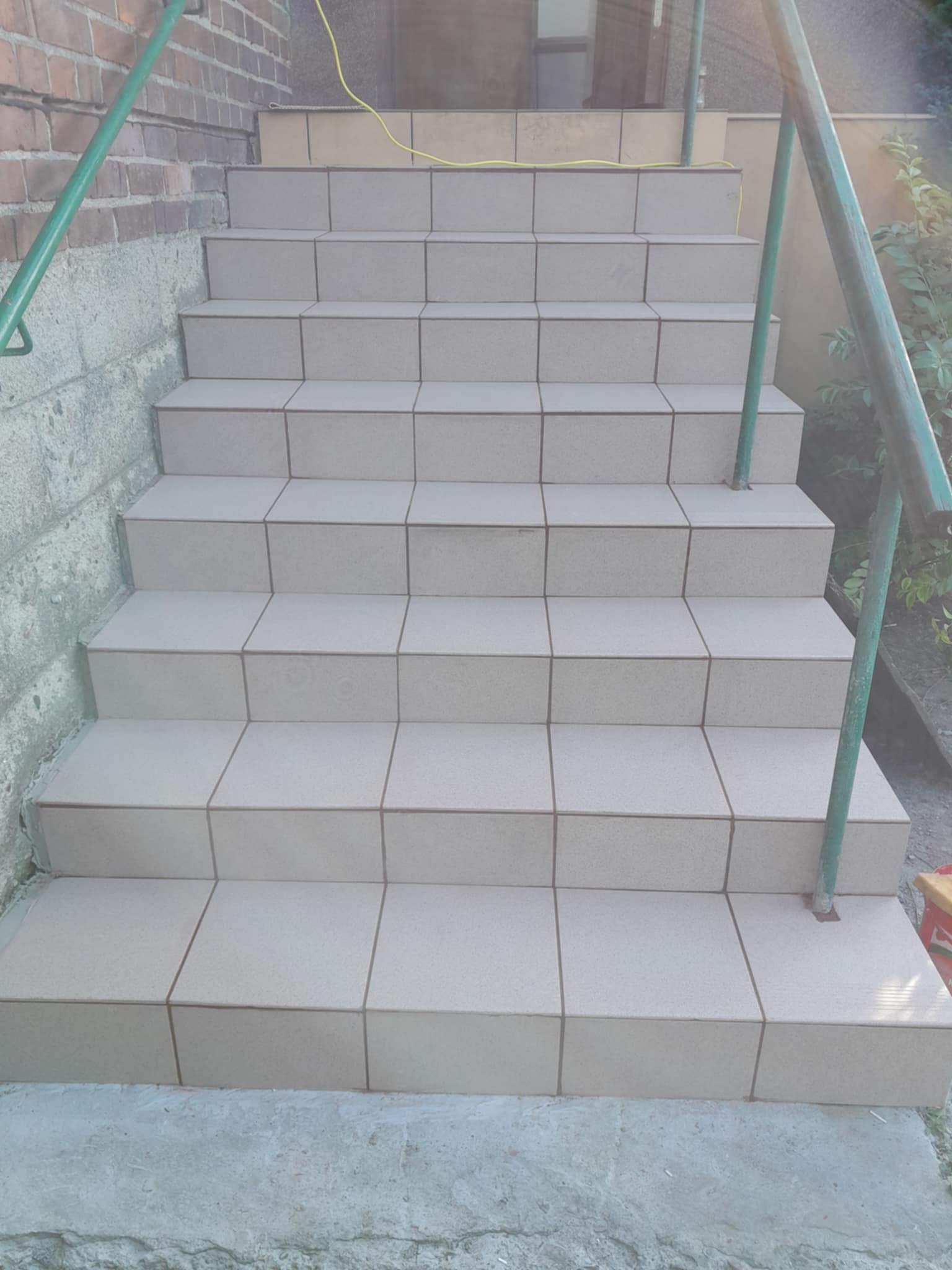 Wykończone schody zewnętrzne z jasnych, kwadratowych płytek, z zieloną metalową barierką po bokach. Widoczne detale fug i struktura płytek.