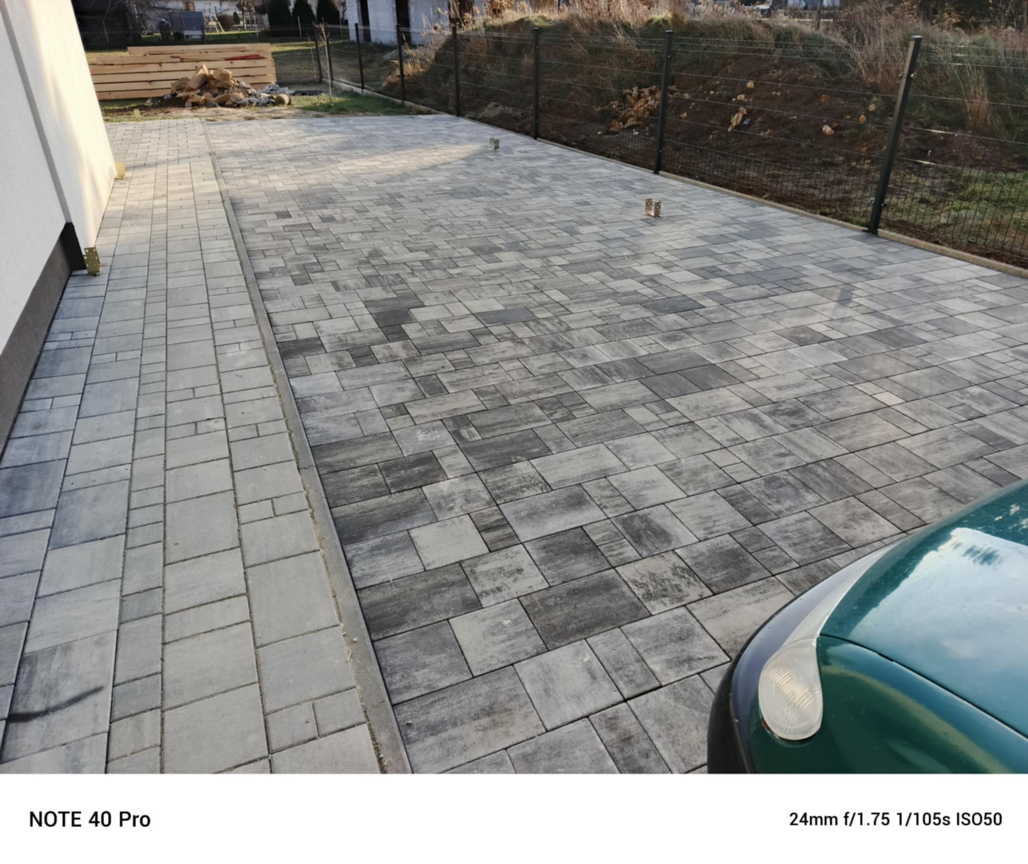 Ułożona kostka brukowa w odcieniach szarości, tworząca wzór na podjeździe. Widoczne obrzeża, fragment ogrodzenia i zaparkowany samochód. Ujęcie z perspektywy.