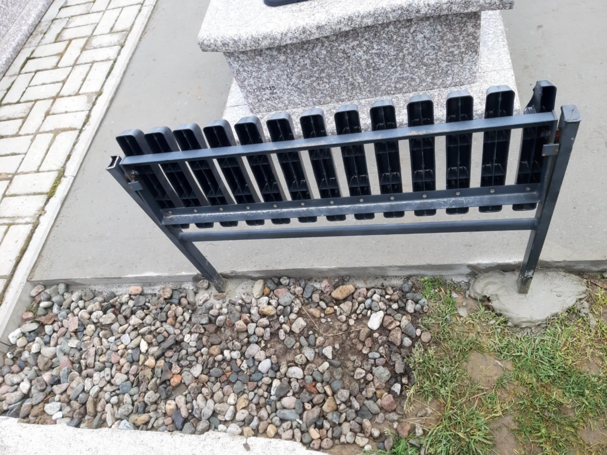 Metalowe ogrodzenie nagrobka, osadzone w betonie, z kamieniami i trawą w tle. Widoczny fragment nagrobka z jasnego kamienia.