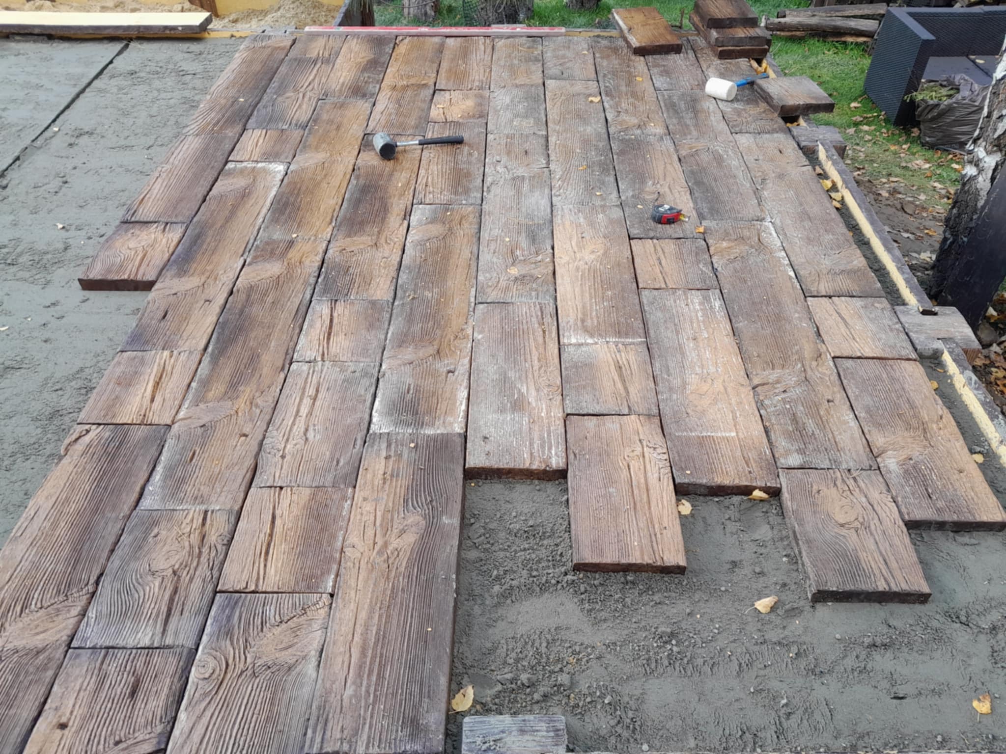 Układanie kostki brukowej imitującej drewno na podsypce piaskowej. Widoczny młotek gumowy i miarka. Częściowo ułożona nawierzchnia z wyraźnym wzorem słojów.
