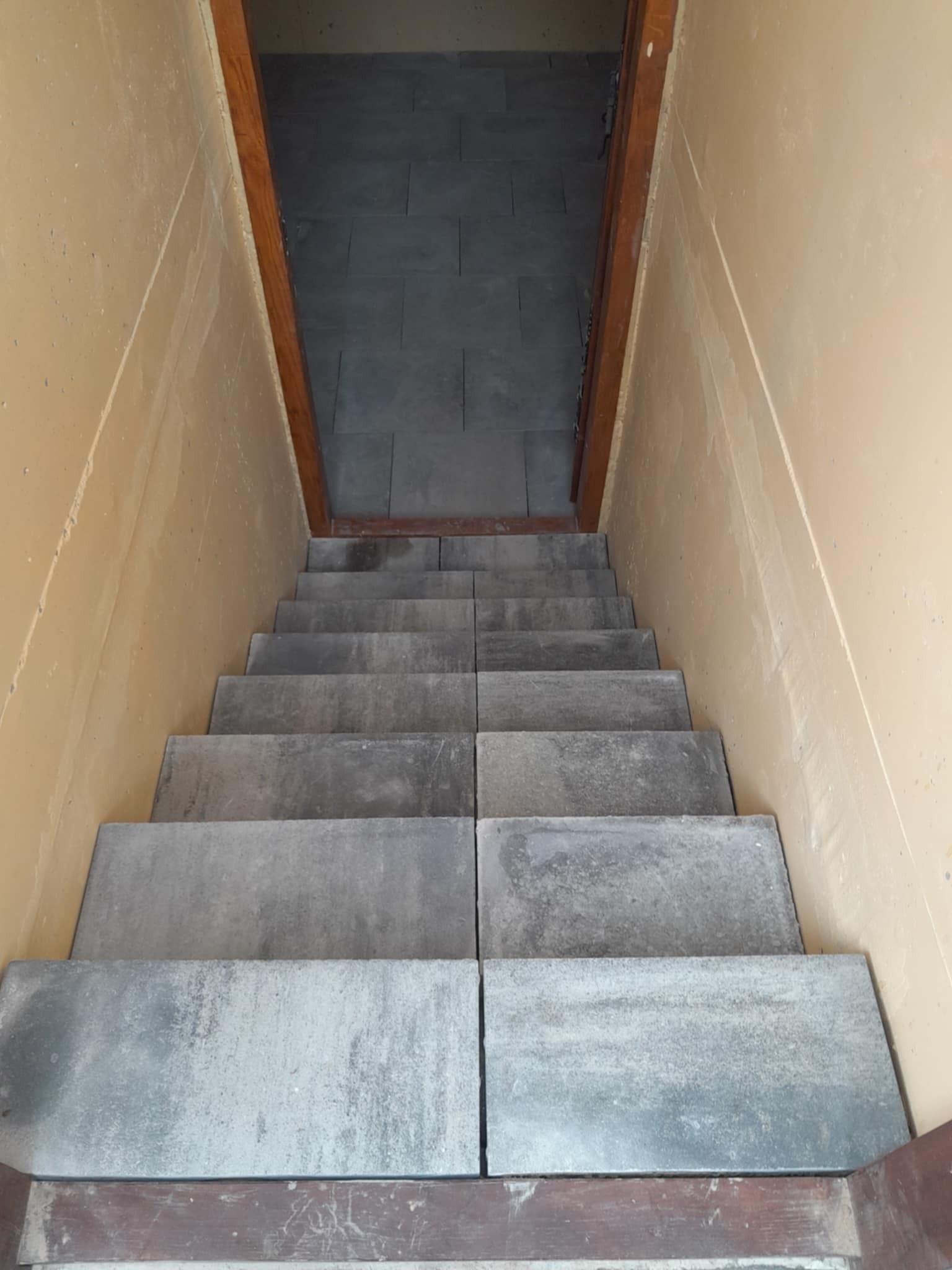 Schody wyłożone szarą kostką brukową, widok z góry. Ściany w kolorze beżowym, drewniana rama drzwi. Kostka ułożona równo, tworząc estetyczne przejście.
