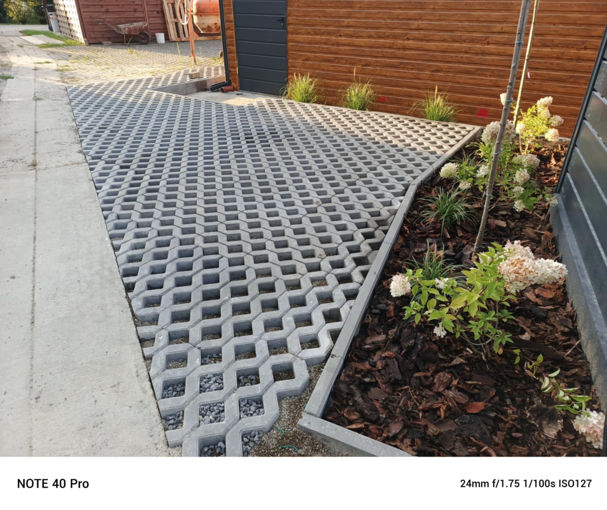 Szara ażurowa kostka brukowa ułożona na podjeździe, wypełniona drobnym kamieniem. Obok rabata z korą i kwitnącymi hortensjami. Drewniana ściana budynku w tle.
