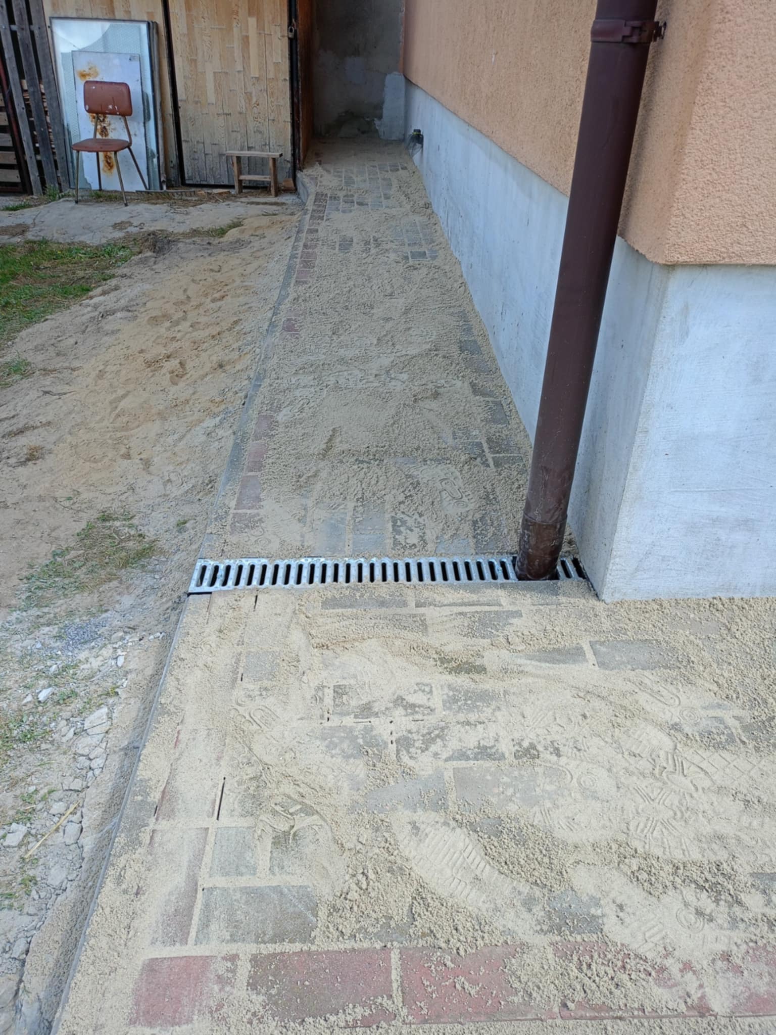 Ścieżka z kostki brukowej w trakcie budowy, zasypana piaskiem, z odpływem liniowym przy ścianie budynku. Widoczne ślady butów na piasku.