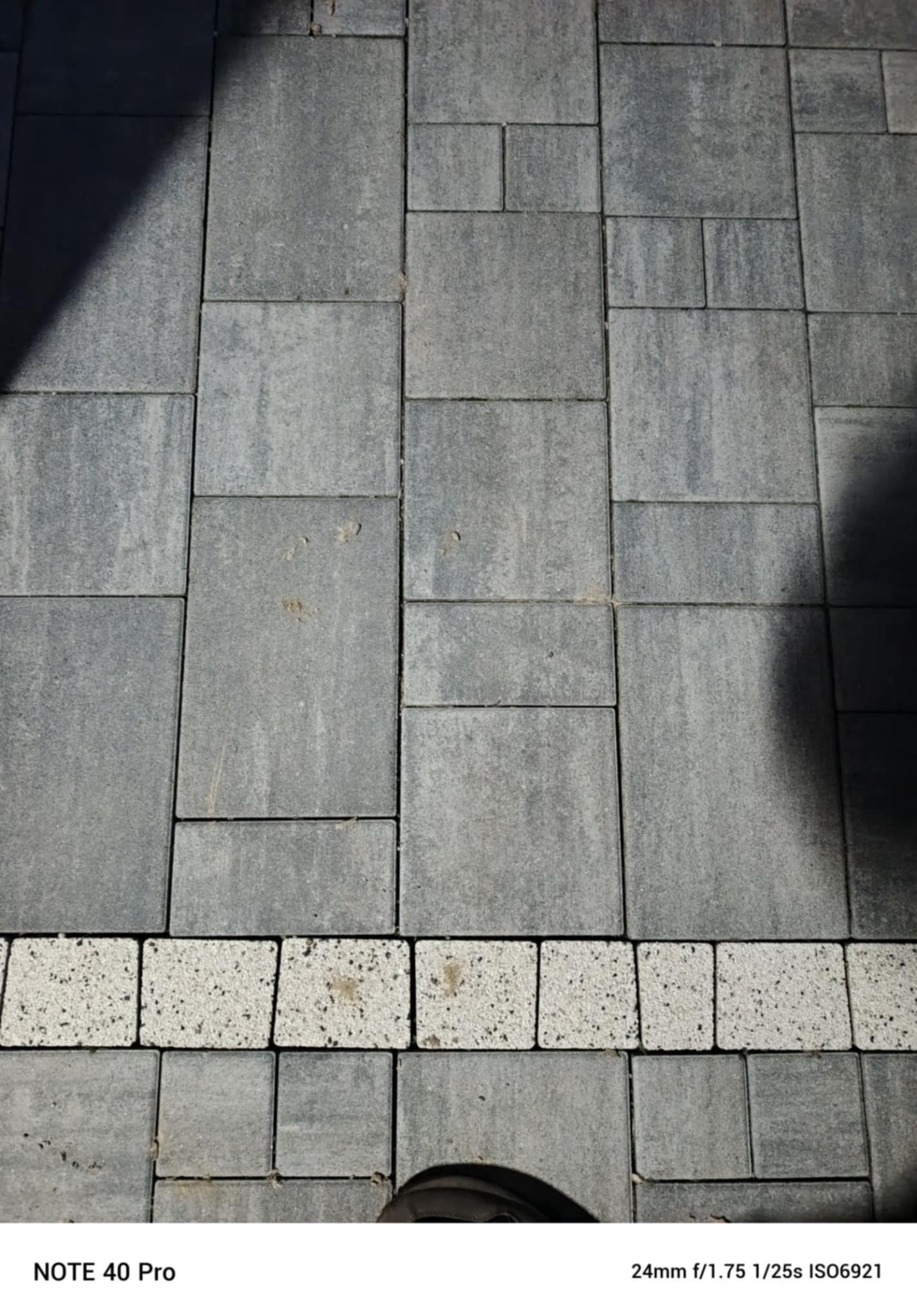 Szara kostka brukowa ułożona w regularny wzór z białym pasem kostki dekoracyjnej. Widoczny fragment buta w dolnej części kadru. Cień częściowo zasłania nawierzchnię.