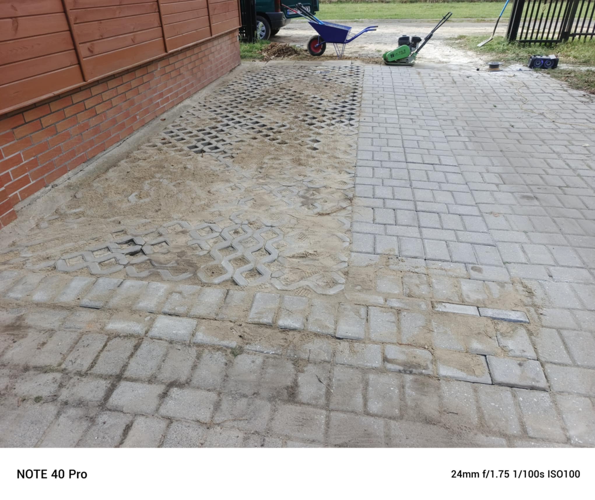 Fragment układanej nawierzchni z kostki brukowej i ażurowej, widoczna ubijarka, taczka i narzędzia. Część kostki zasypana piaskiem, etap prac brukarskich.