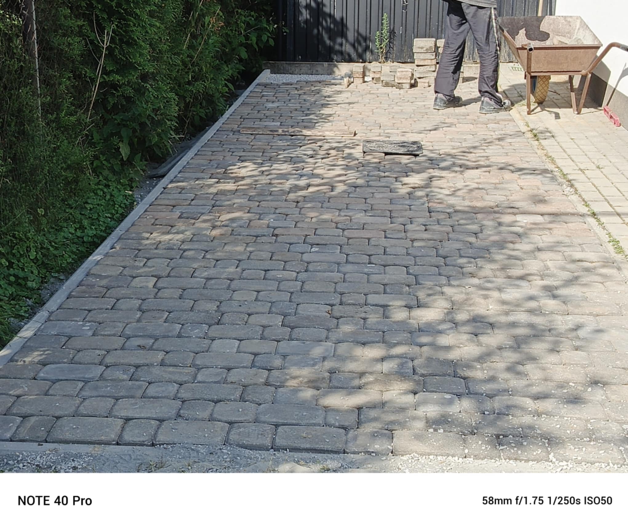Ułożona kostka brukowa w odcieniach szarości, widoczna taczka i fragment osoby w tle, obrzeża z kostki. Częściowo zacieniona powierzchnia.