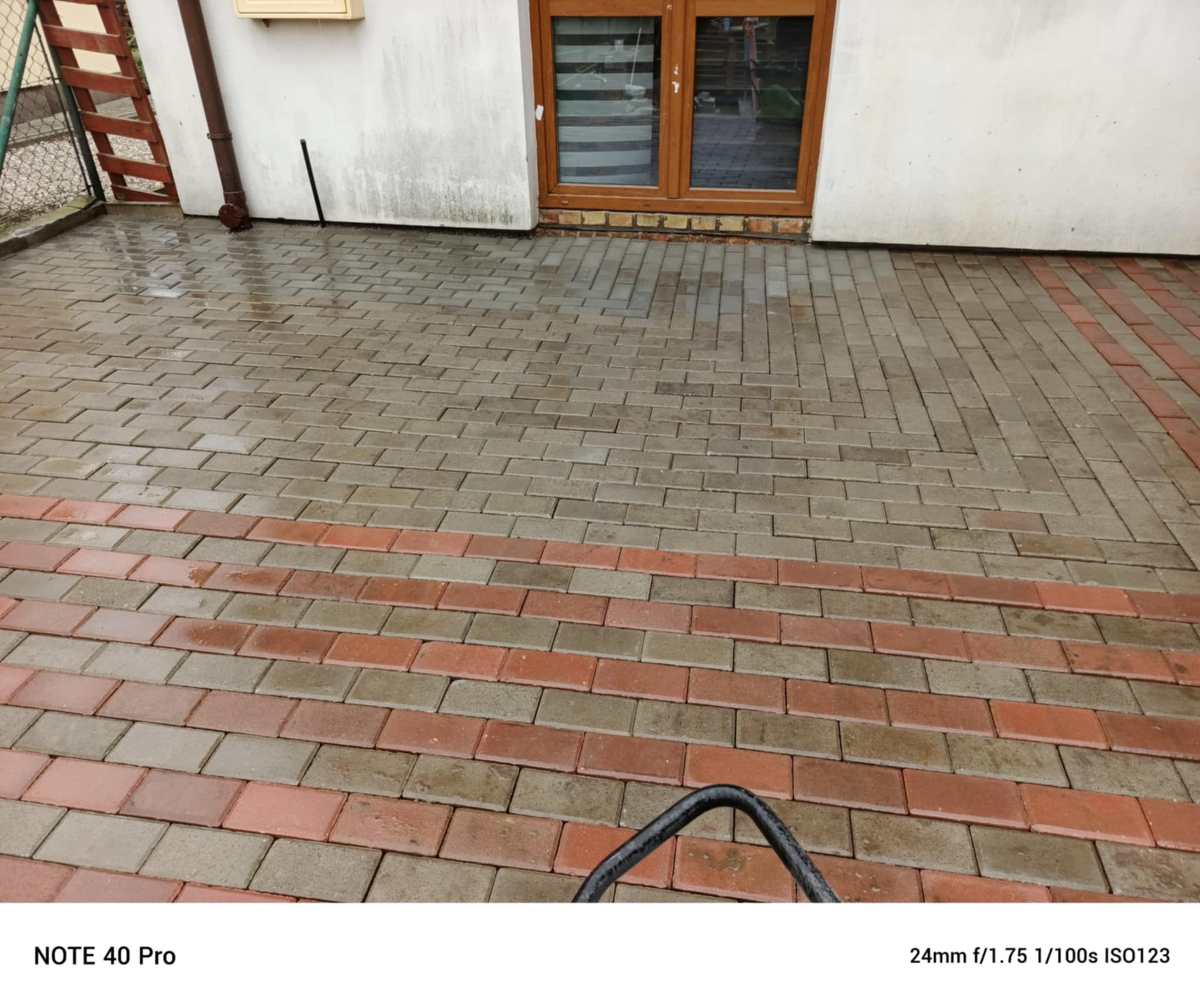 Ułożona kostka brukowa w odcieniach szarości i czerwieni, tworząca wzór na podjeździe. Widoczne fragmenty elewacji budynku i okna. Świeżo po deszczu, mokra nawierzchnia.