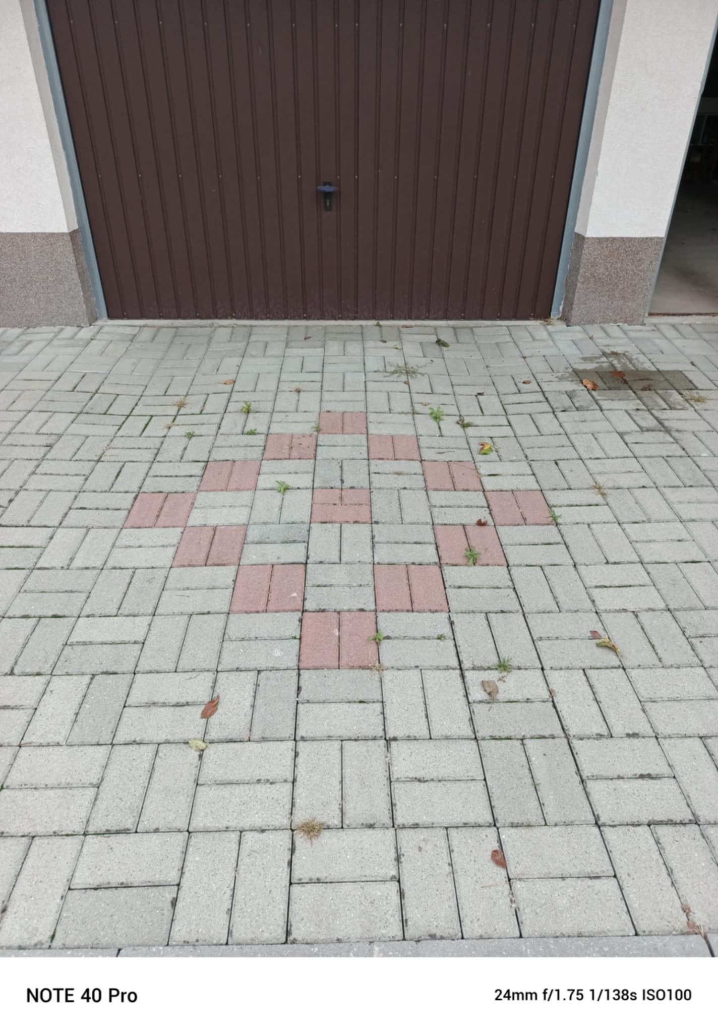 Szara kostka brukowa z czerwonym wzorem krzyża przed brązową bramą garażową. Widoczne drobne chwasty wyrastające spomiędzy kostek. Ujęcie z bliska, perspektywa z góry.