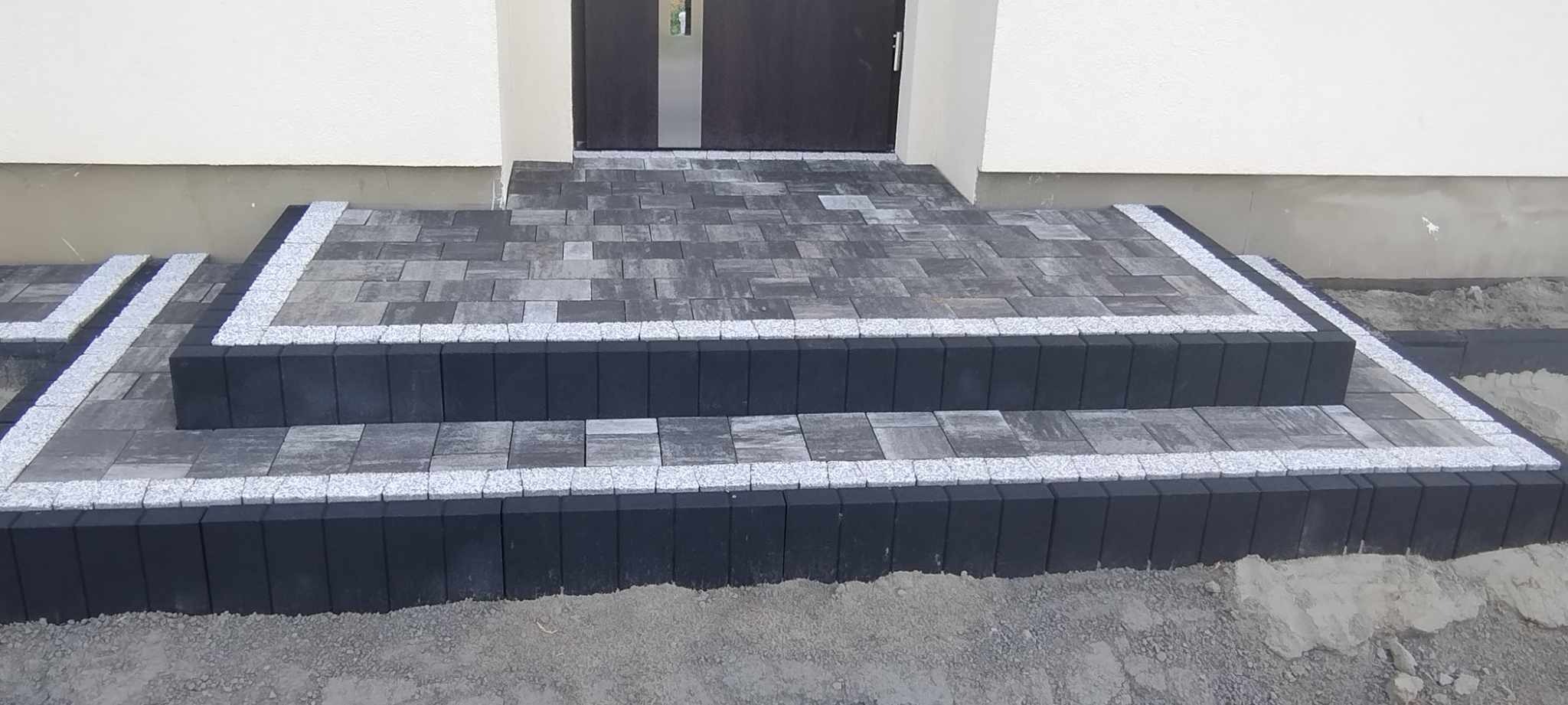 Schody wejściowe wyłożone szarą kostką brukową z obramowaniem z jasnej kostki granitowej, prowadzące do ciemnych drzwi wejściowych.