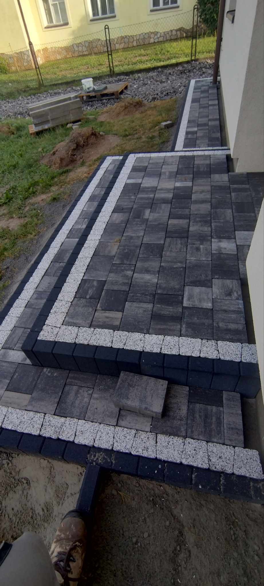 Ułożona kostka brukowa w odcieniach szarości z obramowaniem z białego grysu i czarnych kostek, prowadząca do budynku. Widoczne zapasy kostki brukowej.