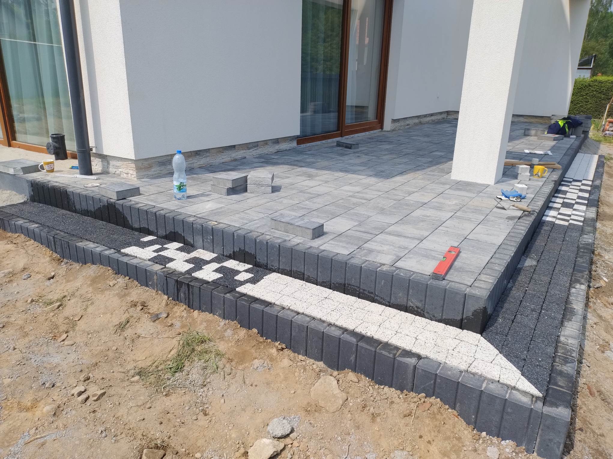 Układanie szarej kostki brukowej na tarasie domu, z dekoracyjnym obrzeżem w czarno-białą szachownicę. Widoczny poziomica i butelka wody. Częściowo ukończony etap prac.